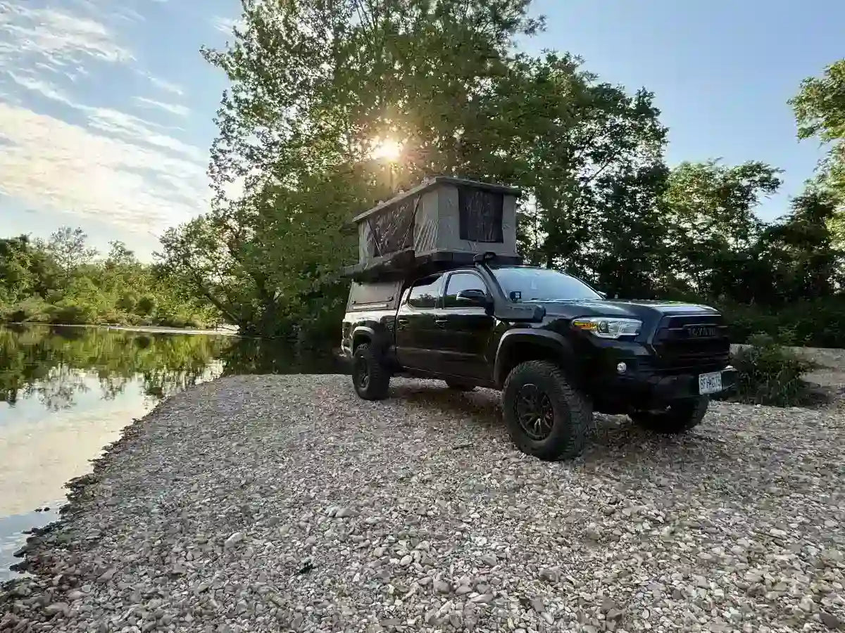 Black Toyota Tacoma with rooftop camper tent parked by a riverbank featuring practical truck camper accessories for outdoor travel and camping.