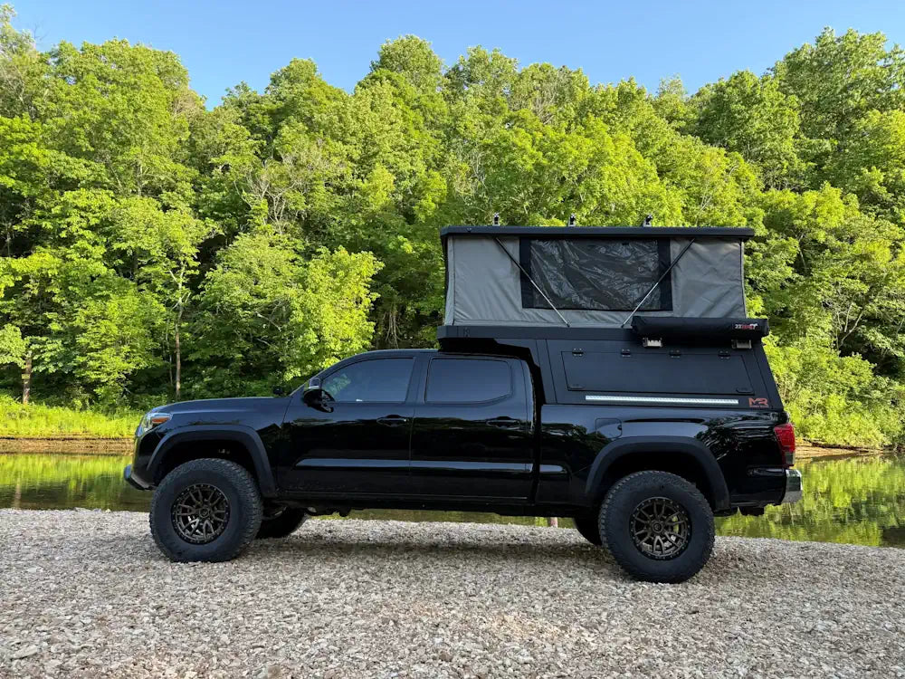 Black truck with pop-up camper parked beside a scenic riverbank, representing a featured highlight from a comprehensive truck camper buying guide.