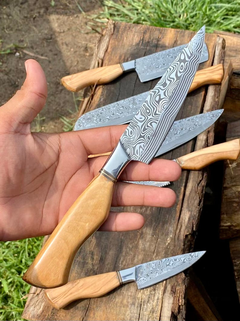 Hand holding a set of Damascus steel knives with wooden handles on a wooden block outdoors.
